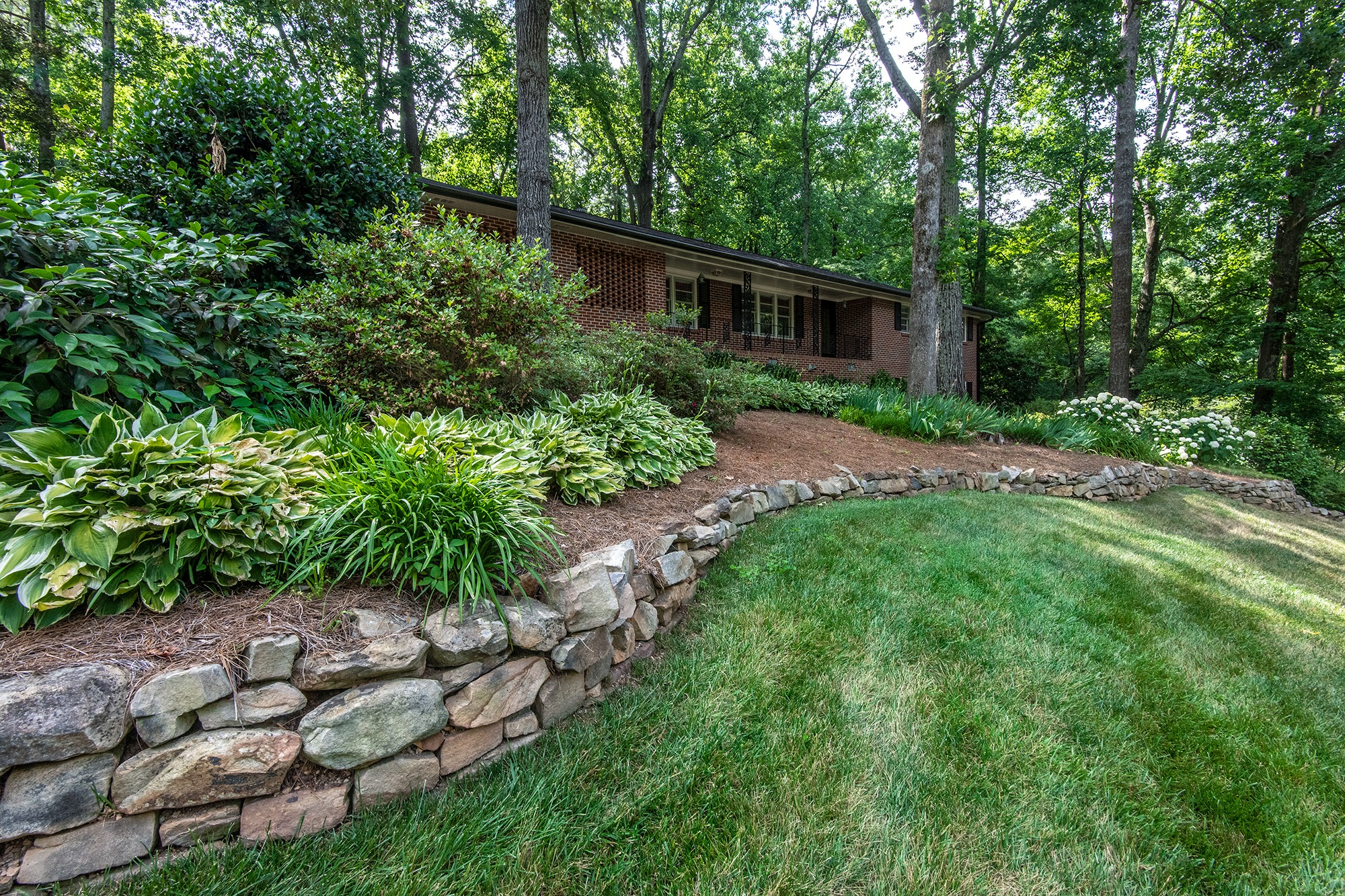 A red brick house is surrounded by lush greenery, stone landscaping, and a well-maintained lawn in a wooded area.