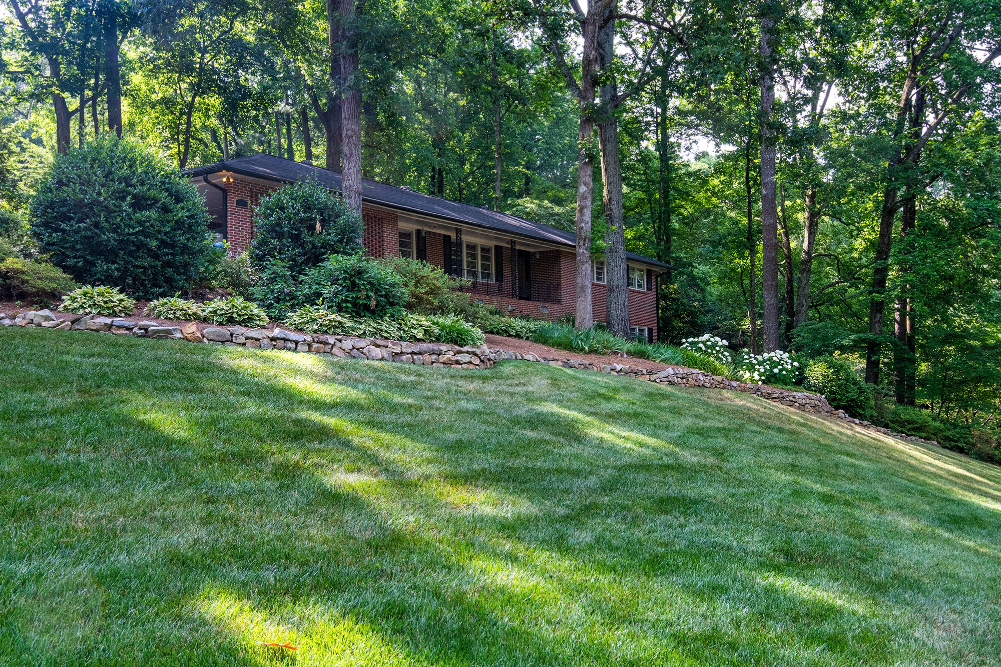 A brick house nestled among tall trees and lush greenery, with a well-manicured lawn and a stone-bordered garden path.