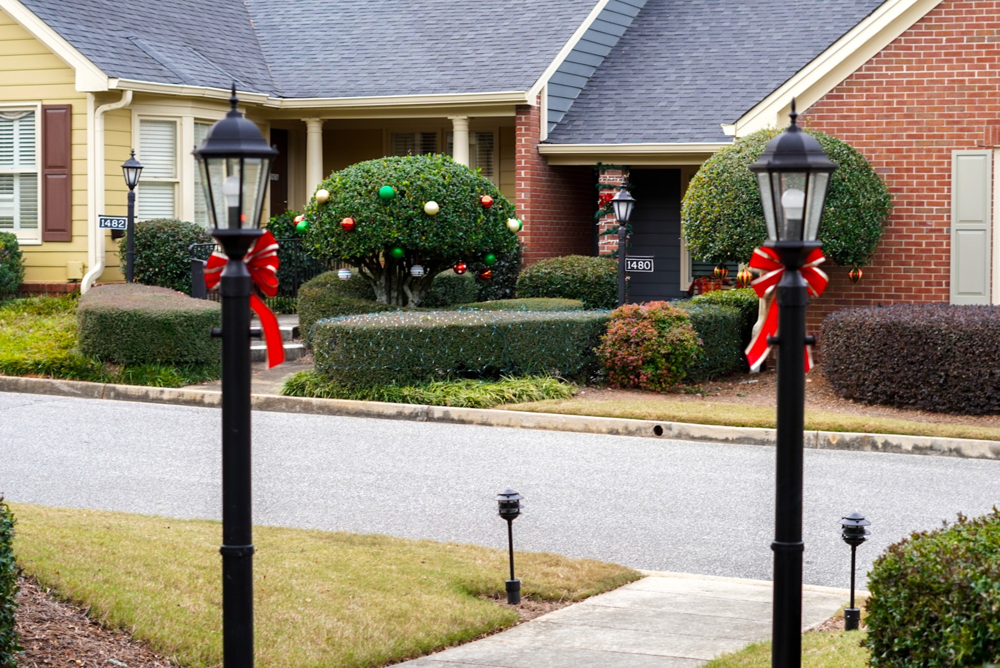 Two decorated lampposts with festive ribbons stand in front of a suburban house and yard adorned with holiday ornaments.