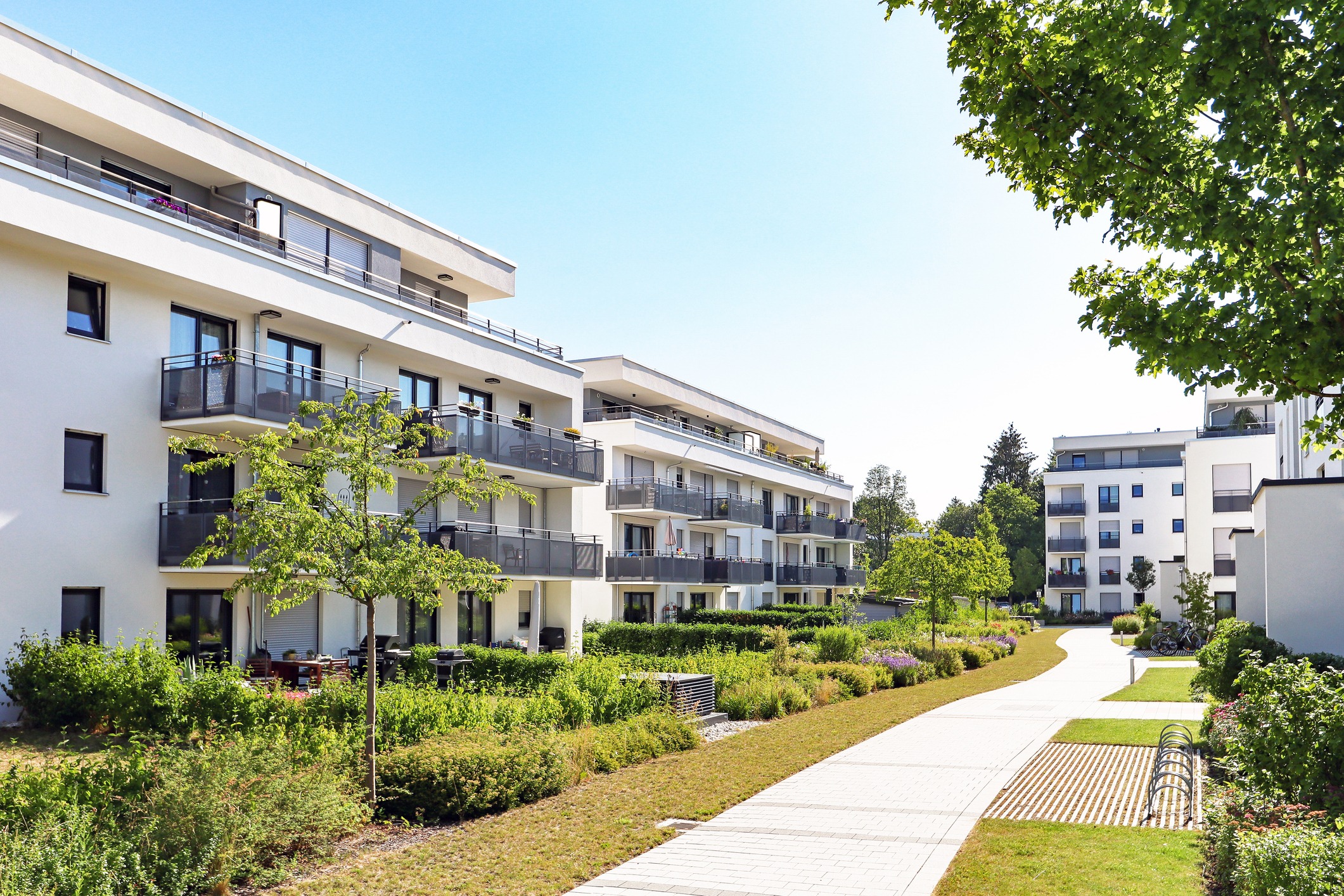 Modern white apartment buildings with balconies, surrounded by lush greenery and pathways, under a clear blue sky on a sunny day.