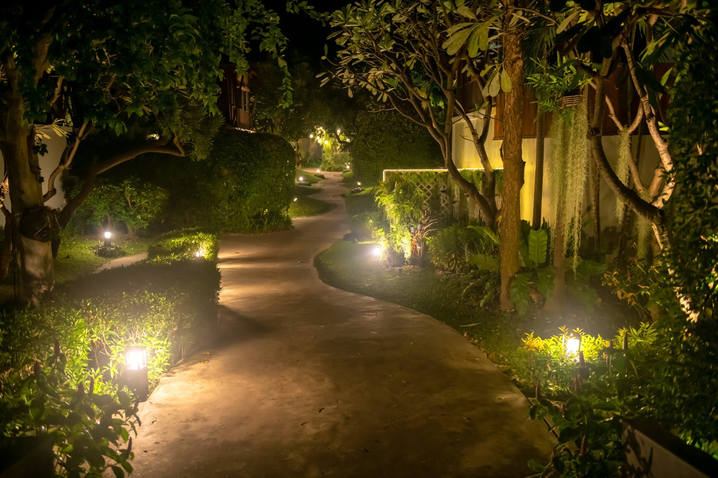 A winding garden path at night, illuminated by small ground lights and surrounded by lush greenery and trees, creating a serene atmosphere.