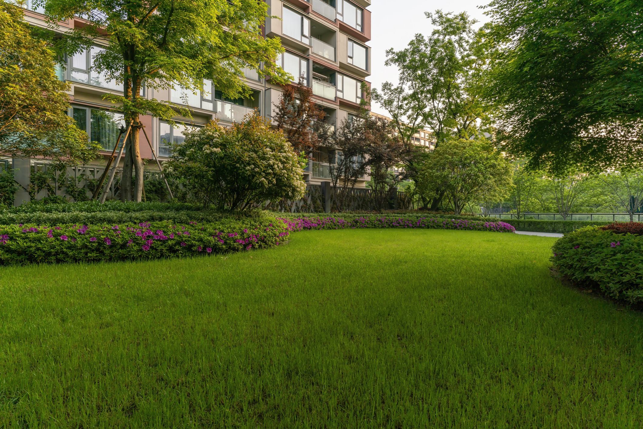 A lush green garden featuring vibrant flowerbeds is nestled next to a modern residential building, surrounded by trees and shrubs.