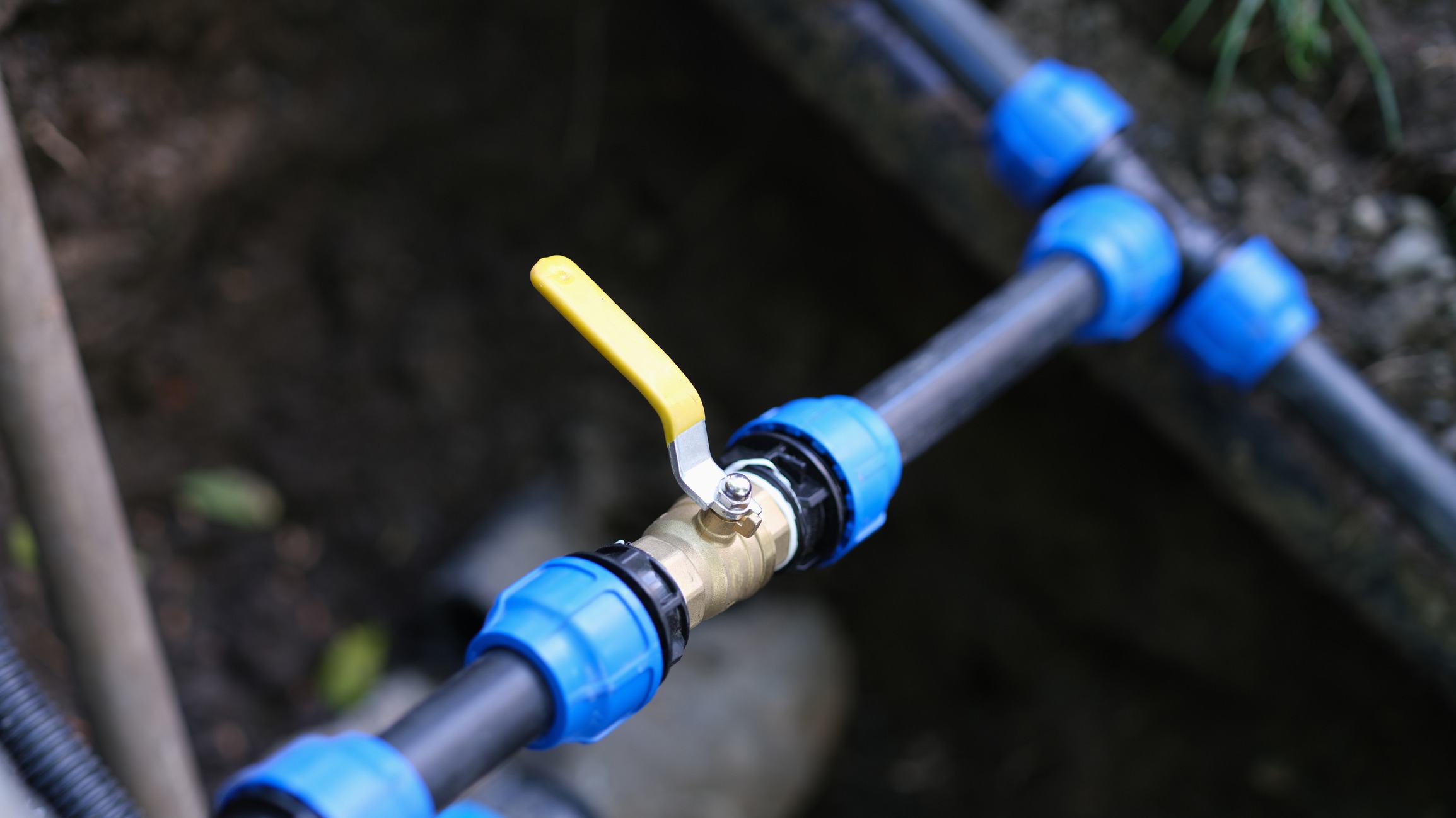 A close-up of a black plastic pipe with a yellow-handled brass valve and blue connectors, outdoors amidst soil and foliage.