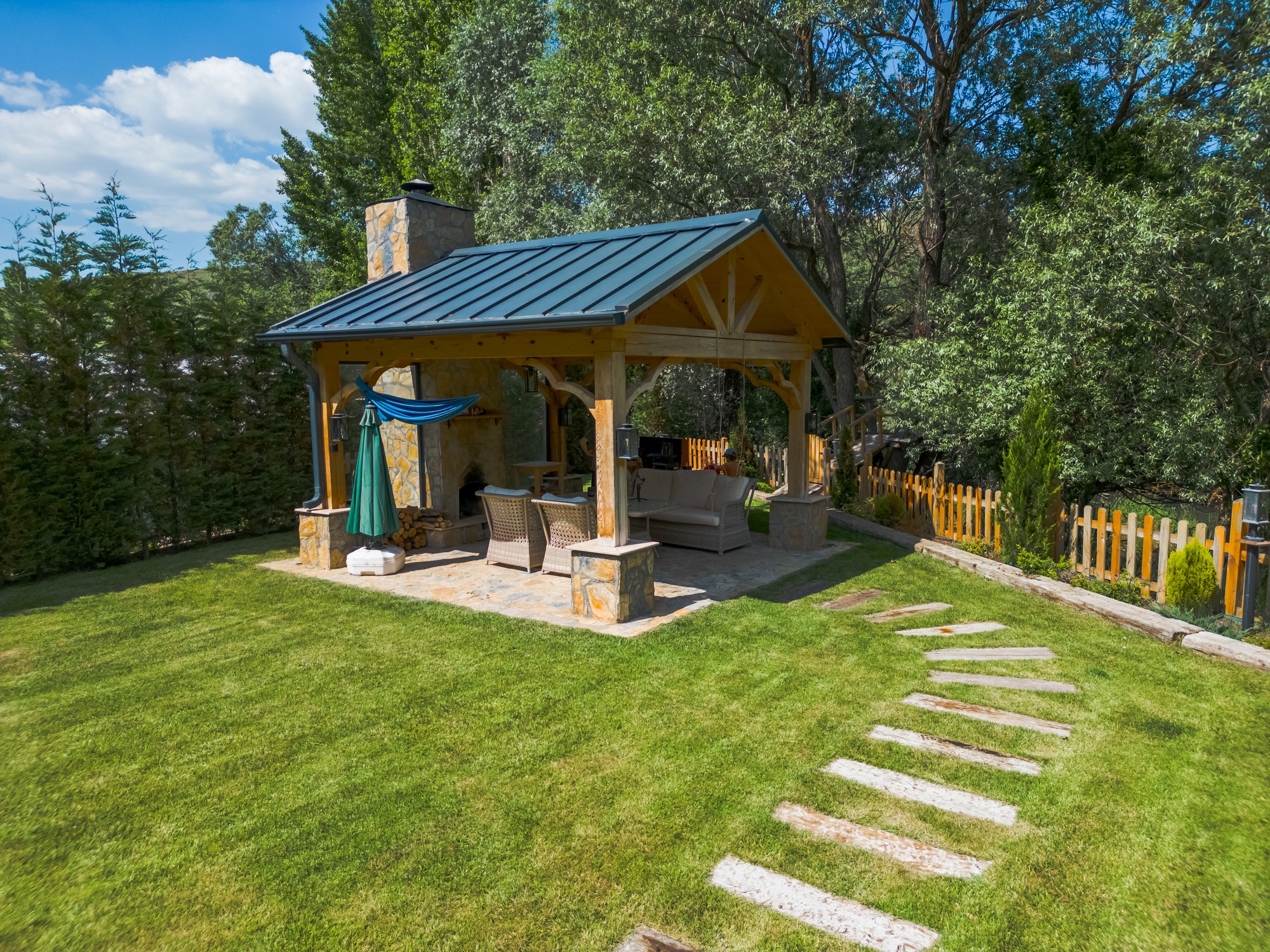 A cozy backyard patio with wooden gazebo, stone fireplace, chairs, umbrella, surrounded by lush grass, trees, and a wooden fence.