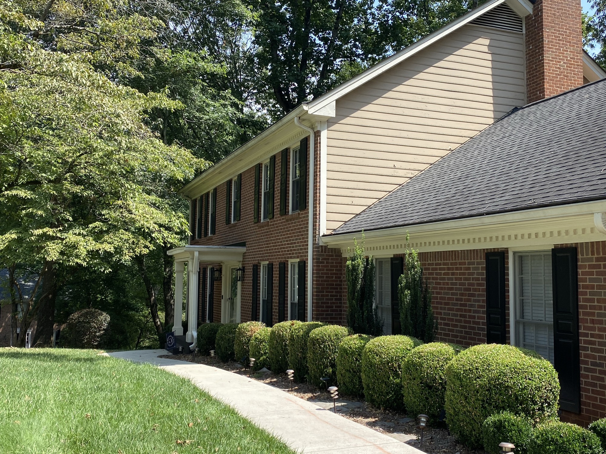 A brick suburban house with black shutters, surrounded by trees and manicured bushes, under a clear sky. No visible landmarks or people.