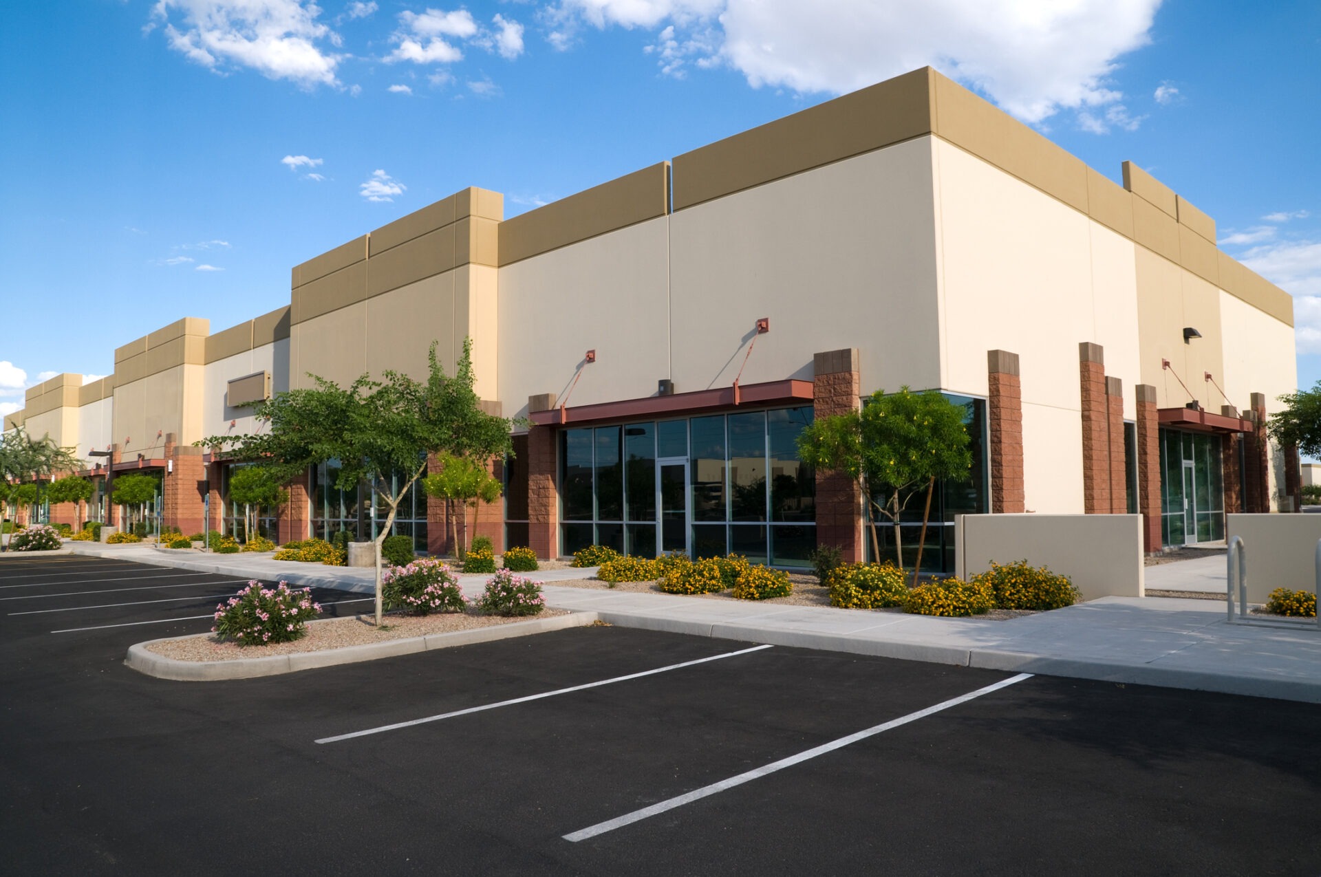 Modern commercial building with glass windows, surrounded by landscaped greenery and an empty parking lot under a clear blue sky with scattered clouds.