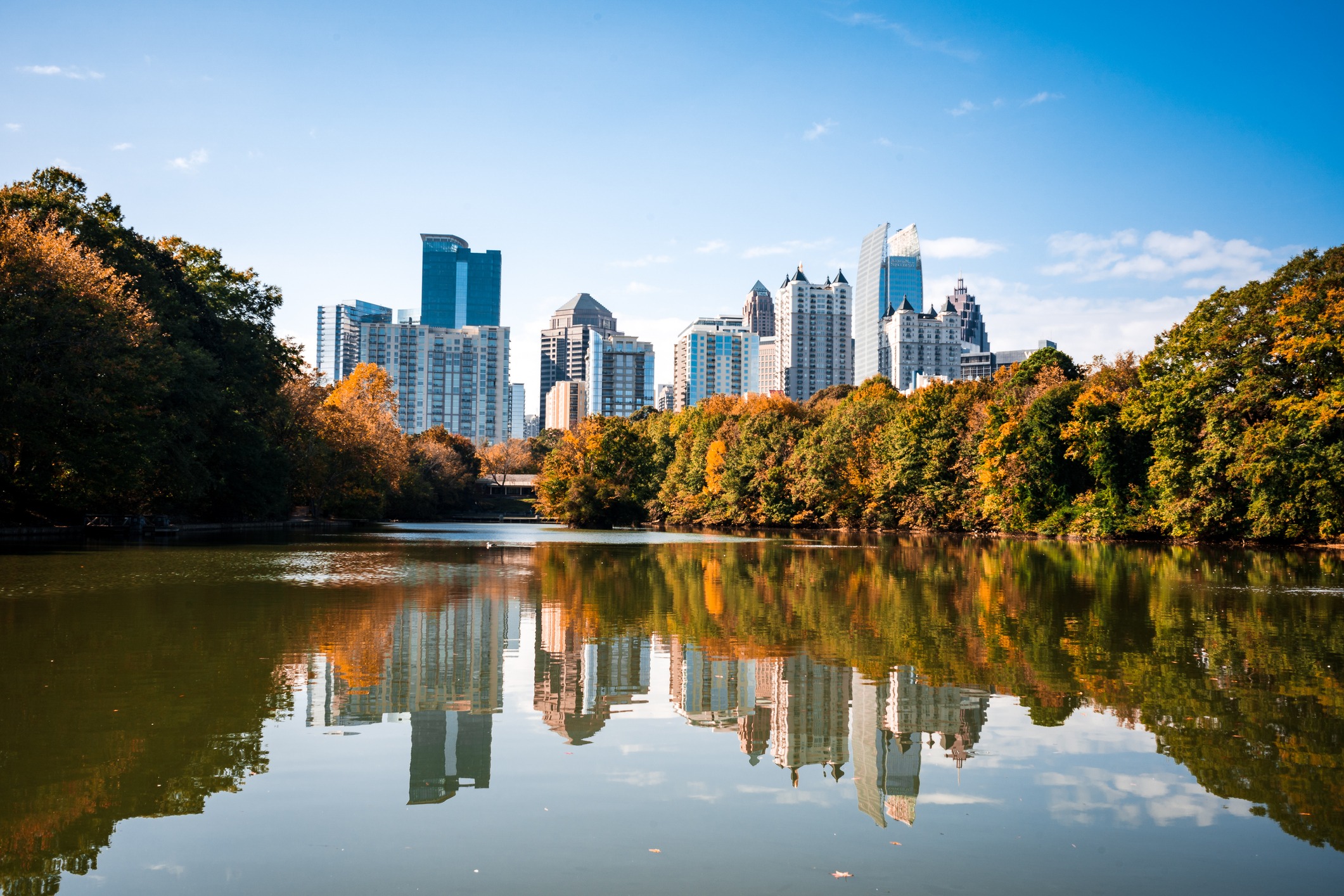 Skyline of Atlanta reflected in a serene lake, surrounded by autumn trees in Piedmont Park, under a clear blue sky.
