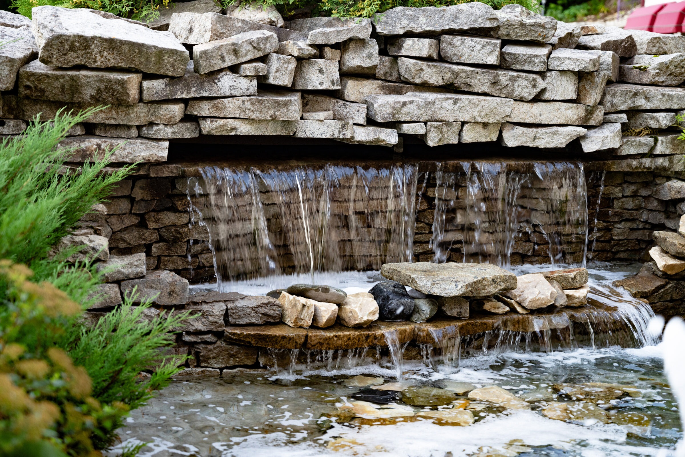 A small waterfall cascades over a rustic stone wall into a pond, surrounded by lush greenery, creating a serene natural setting.