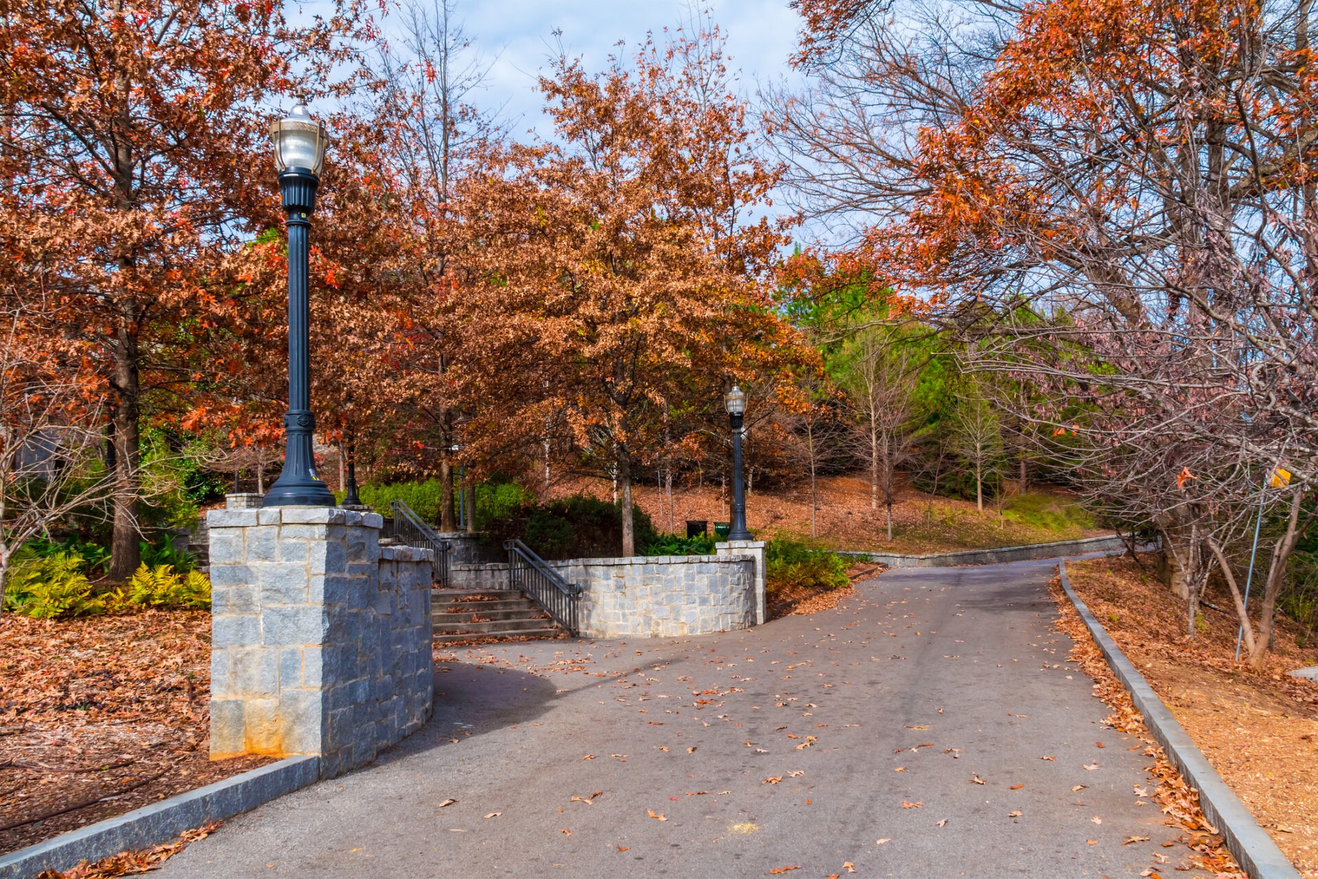 Autumn scene featuring paved path, stone barriers, and lampposts. Trees with orange leaves line the path, creating a peaceful, seasonal atmosphere.