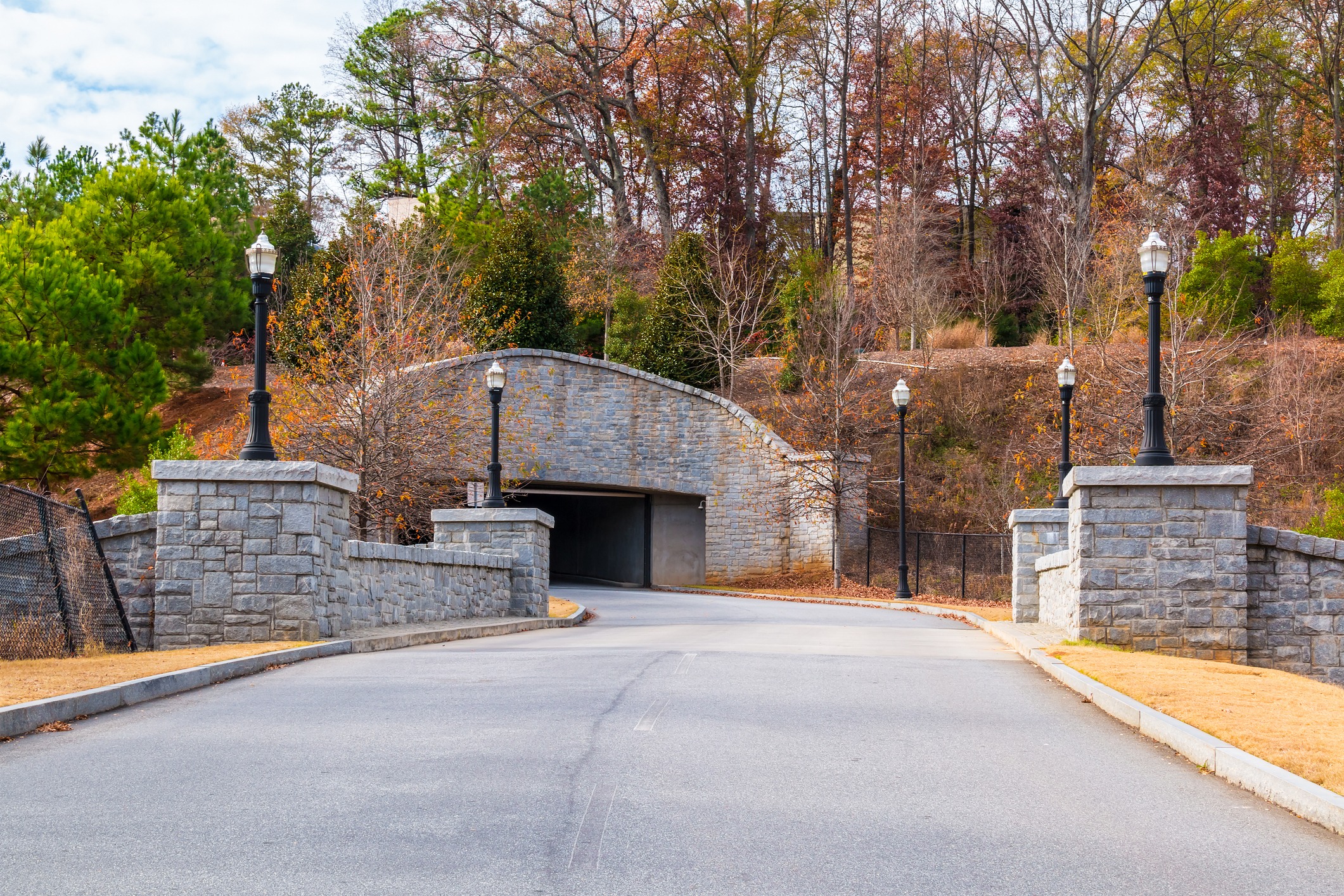 A curved stone bridge with lampposts, surrounded by autumn trees, leading to a tunnel entrance on a quiet, empty road.