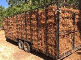 A trailer loaded with hay bales is parked on a dirt path, surrounded by trees under a clear blue sky.