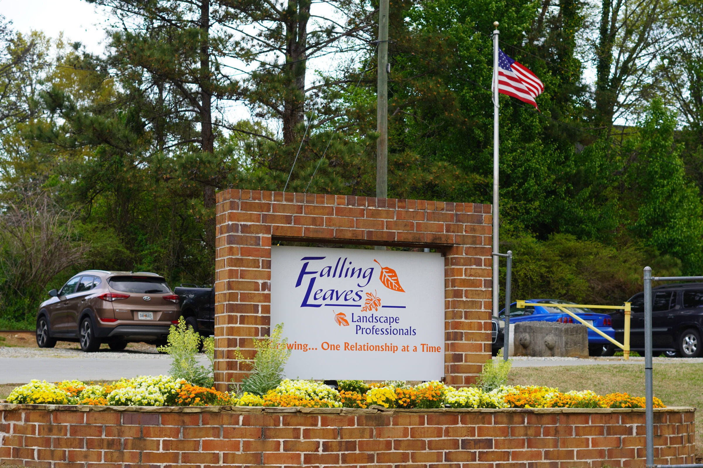 A brick sign for "Falling Leaves Lawn Care" surrounded by colorful flowers, parked cars, and lush greenery in the background.