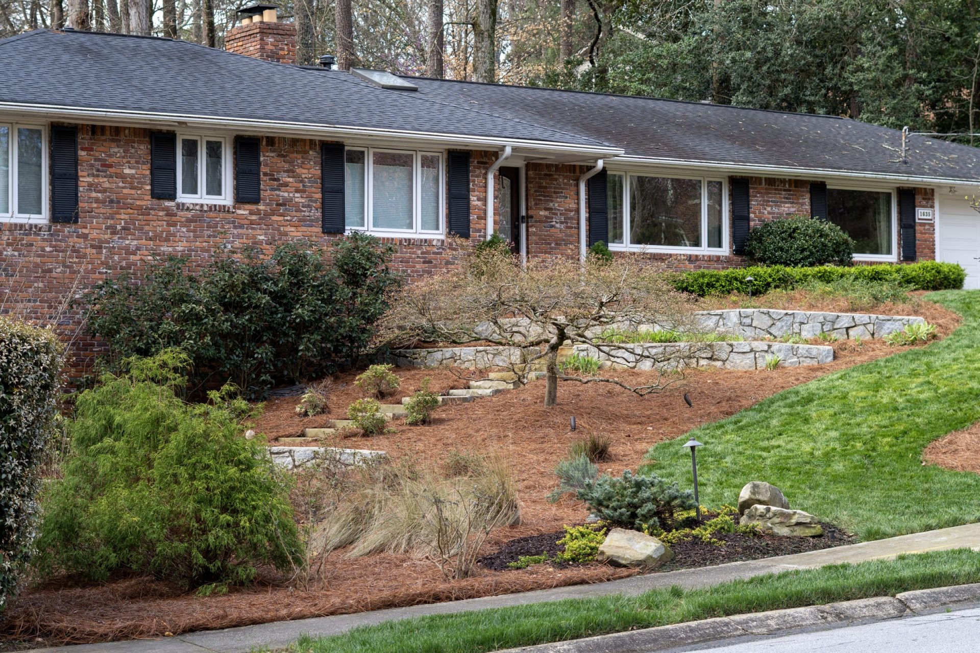 A single-story brick house with a dark shingled roof, featuring a multi-tiered front yard landscape with stone retaining walls, a winding stone staircase, pine straw mulch, a decorative Japanese maple, and a maintained green lawn on the right.