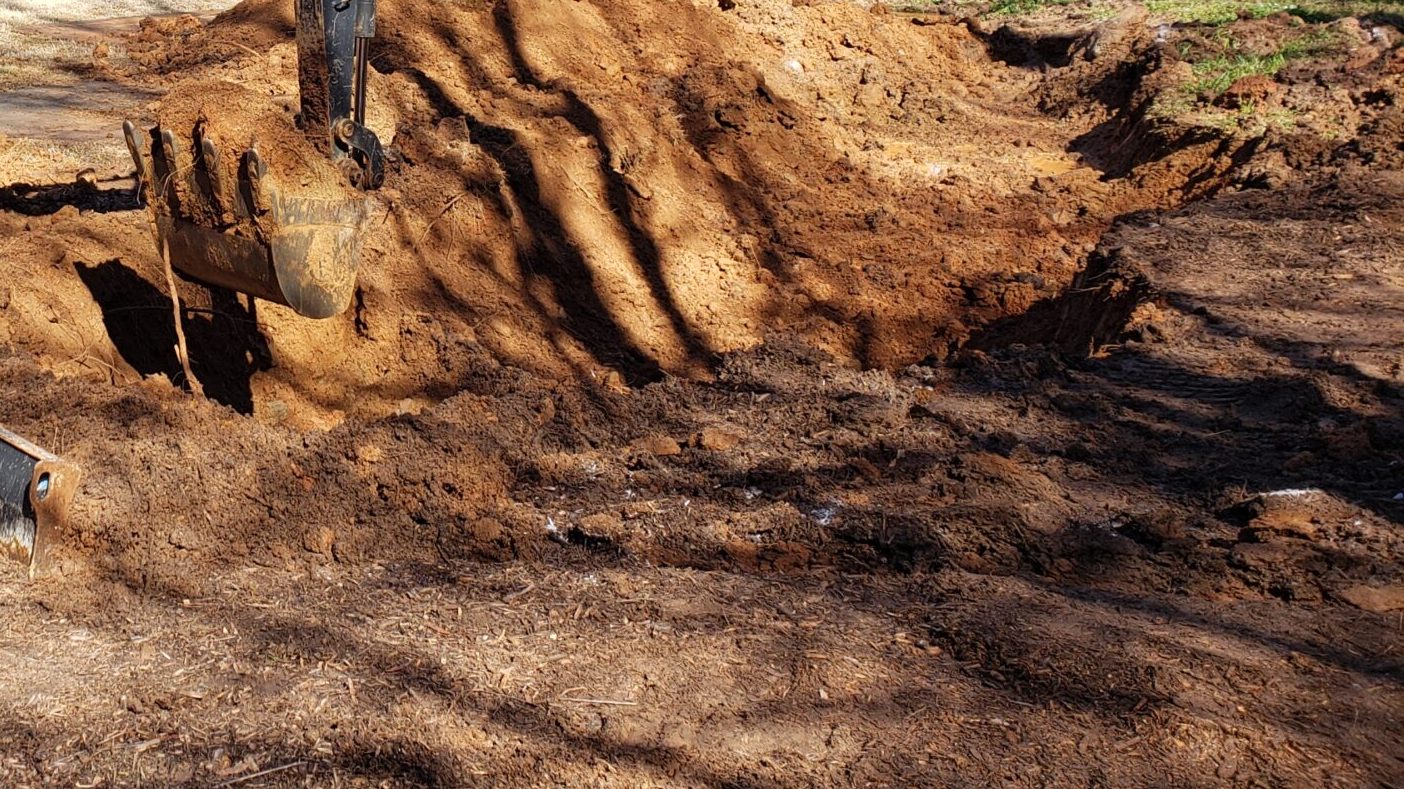 Two persons operate construction equipment, digging soil at a park. They're surrounded by trees and a fence under a clear sky.