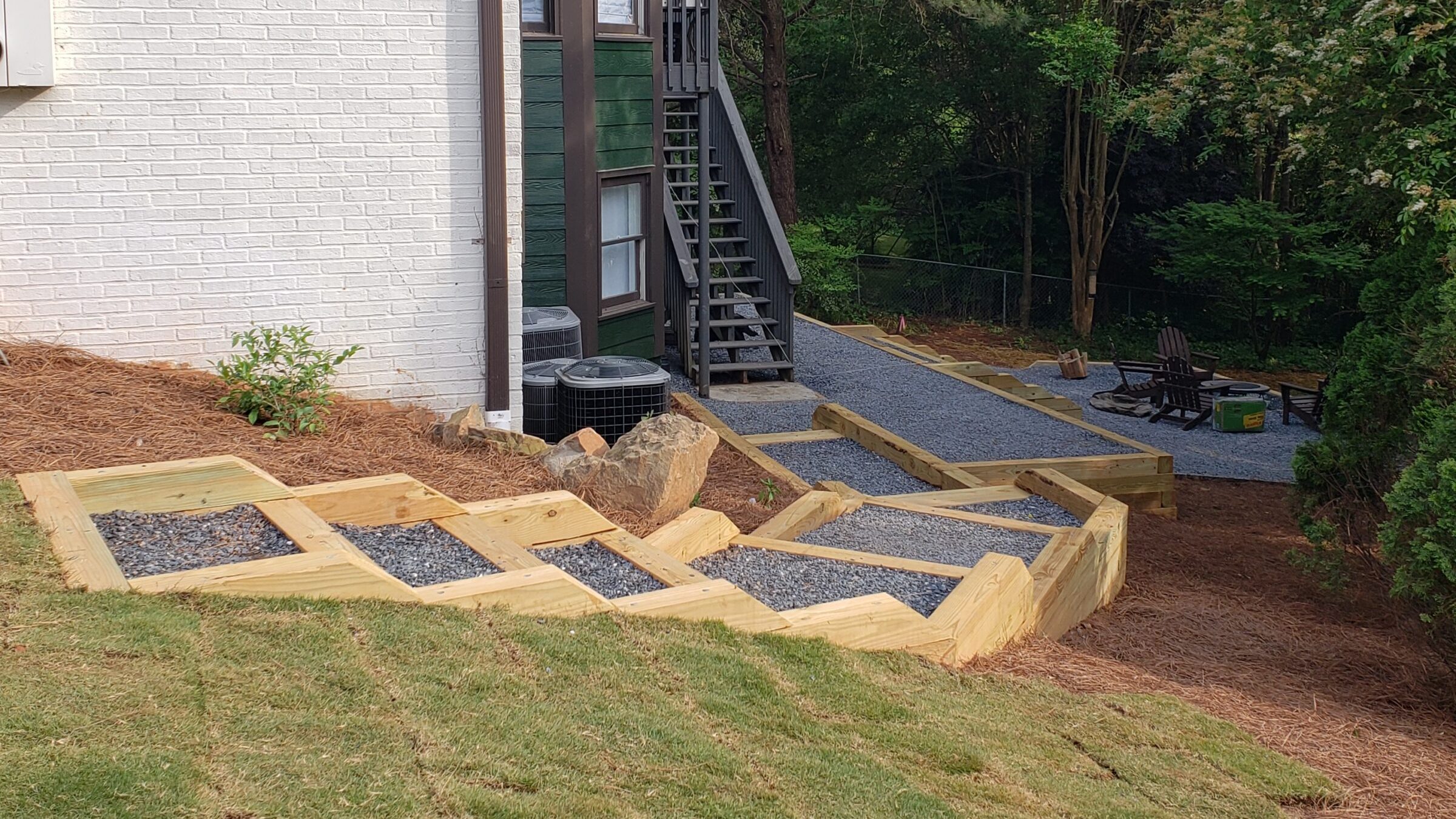 A side yard of a house with white brick and green siding, gravel pathway, wooden steps, surrounded by trees and an outdoor seating area with fire pit.
