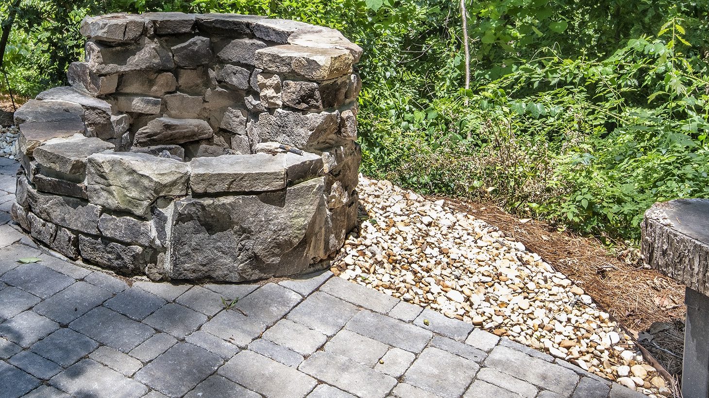 Stone fire pit with curved wooden benches surrounded by lush greenery, placed on a paved patio. Peaceful outdoor setting for relaxation.